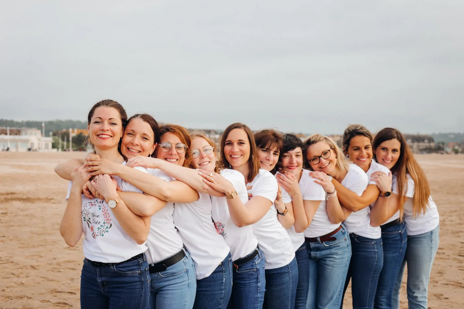 Groupe de dix femmes souriantes qui s'enlacent les unes les autres sur la plage, portant toutes des t-shirts blancs et des jeans, avec un ciel nuageux en arrière-plan.