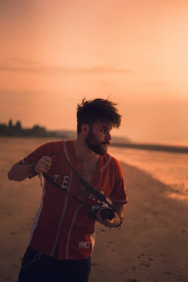 Un homme avec des cheveux bruns, une barbe, portant un t-shirt rouge, tenant un appareil photo à la main, marche sur une plage au coucher du soleil.