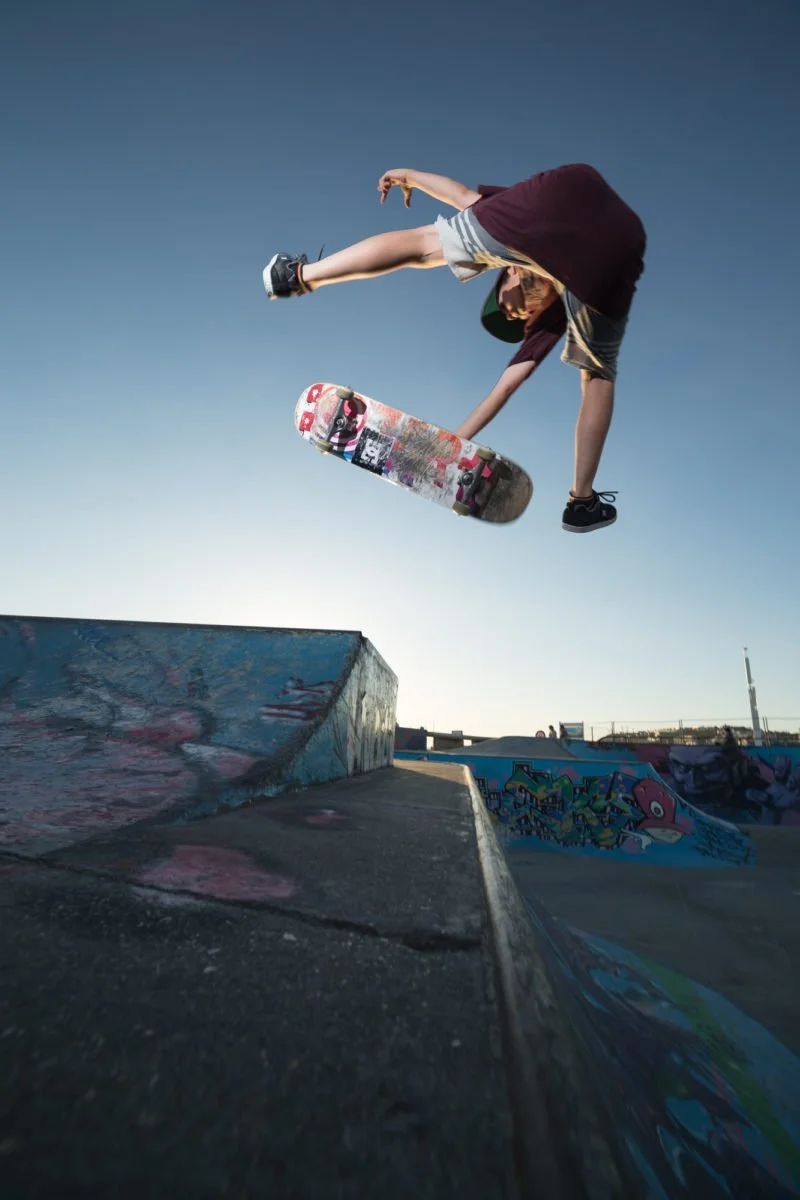 Un jeune garçon en skate en train de faire un saut au-dessus d'une rampe dans un skatepark, avec un ciel clair en arrière-plan.