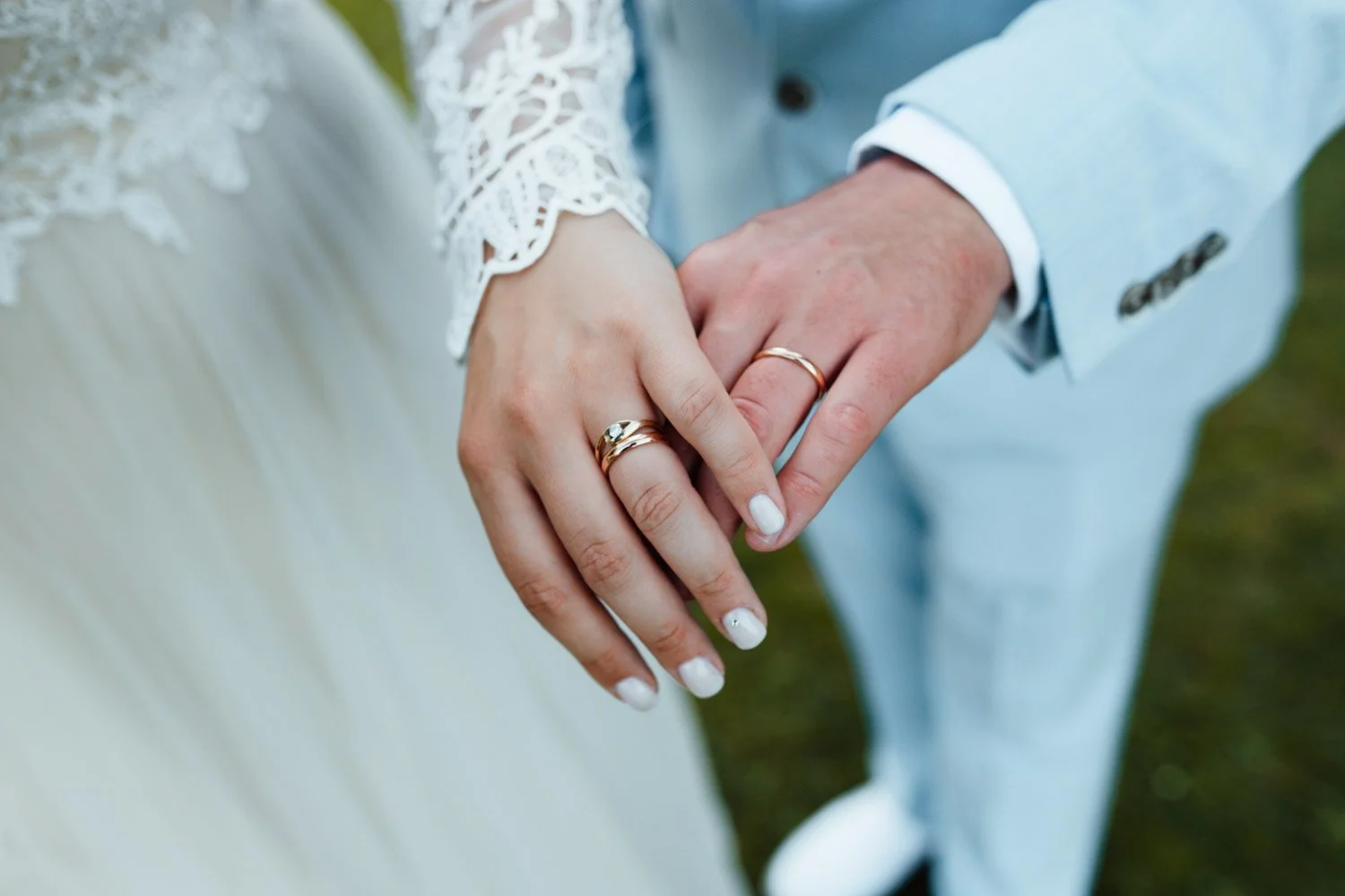 Premier plan de la main d'un couple marié portant des alliances, la femme a des ongles blancs et porte une robe blanche avec des détails en dentelle, l'homme porte un costume bleu clair.