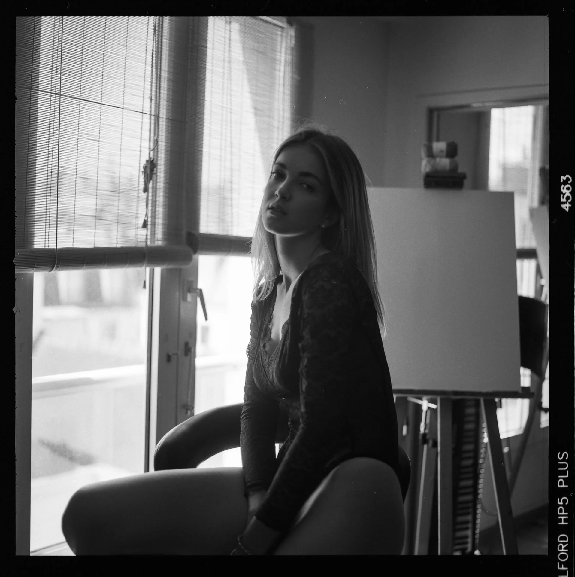Un portrait noir et blanc d'une jeune femme avec des cheveux longs, posant devant une fenêtre avec des stores enroulés. La lumière naturelle illumine une partie de son visage.