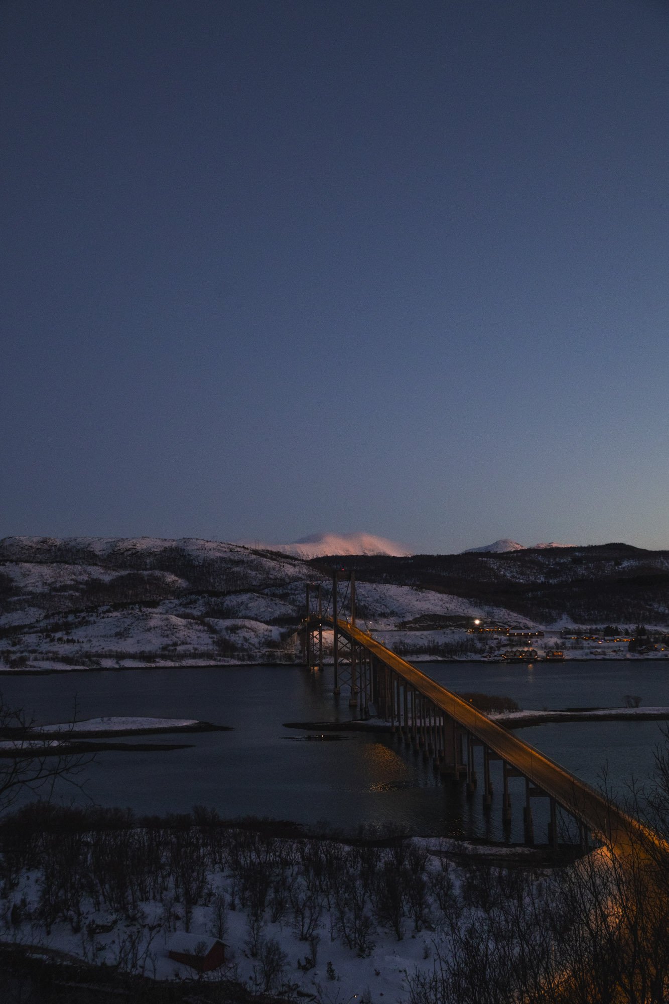 Un pont suspendu en acier long traverse un lac en hiver avec des montagnes enneigées au fond, sous un ciel crépusculaire à Tromsø.