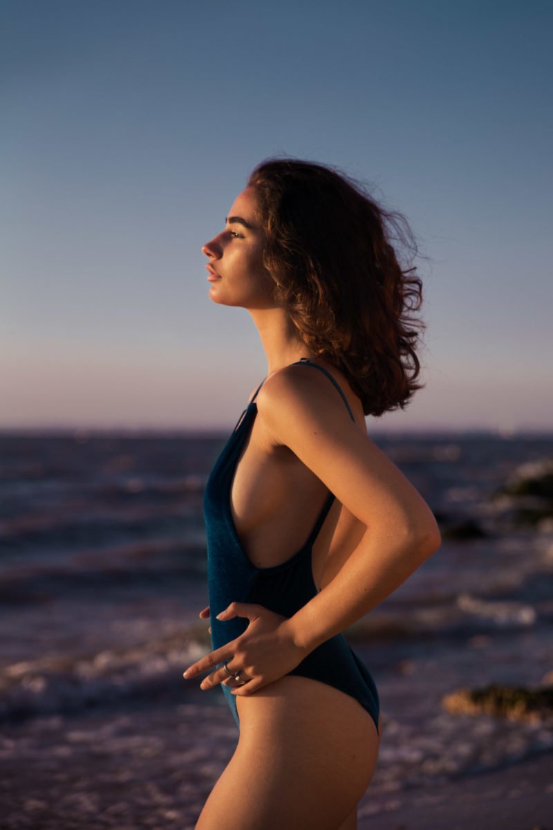 Jeune femme debout au bord de la mer, portant un maillot une pièce bleu, regardant vers l'horizon au coucher du soleil.