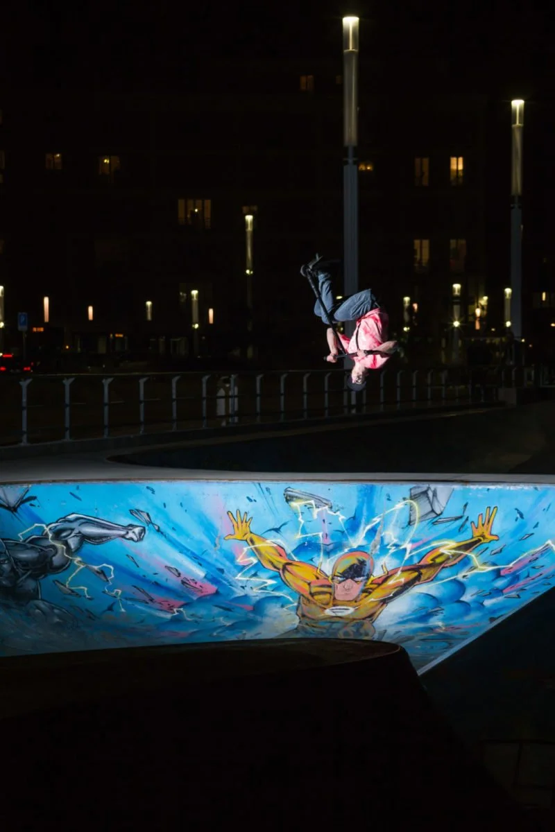 Un garçon en rollerdebout dans un skate-park la nuit, éclairé par des lampadaires, avec une peinture murale de super-héros au fond.