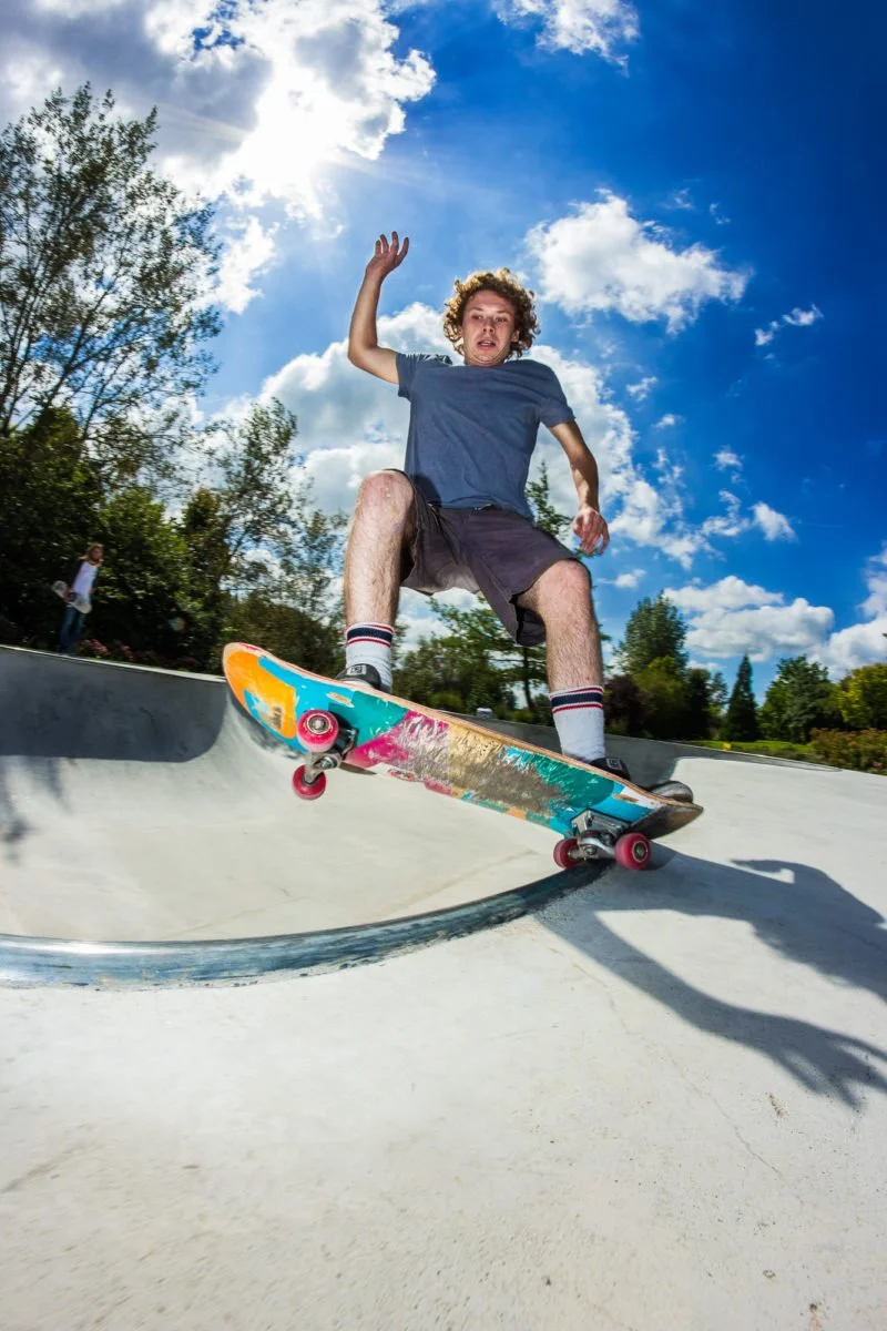 Jeune homme faisant du skateboard dans un skatepark en plein air, soleil brillant et ciel bleu avec quelques nuages.