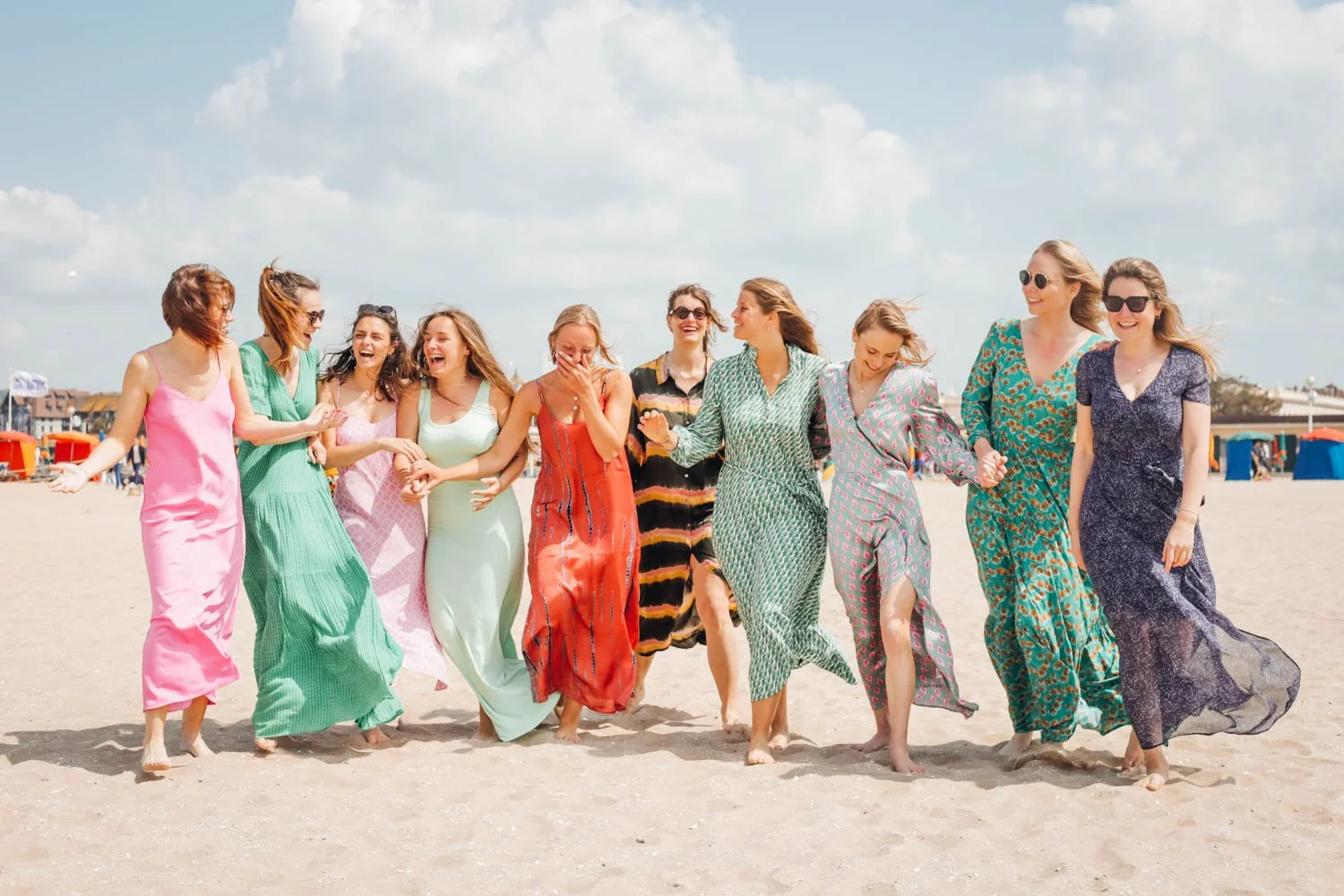 Groupe de femmes souriantes et tenant la main qui marche sur la plage sous un ciel ensoleillé, portant des robes colorées.