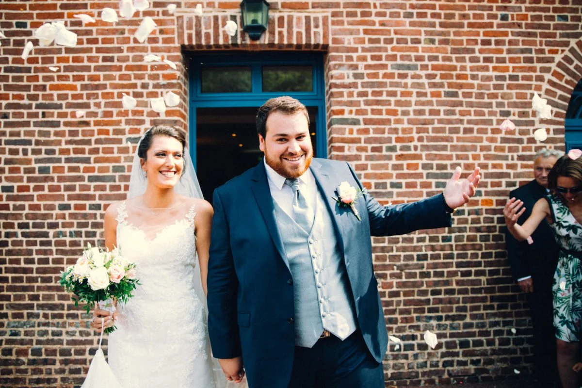 Un couple de mariés souriants sortant d'un bâtiment en briques, entourés de membres de la famille et d'amis, avec des pétales de rose dans l'air.
