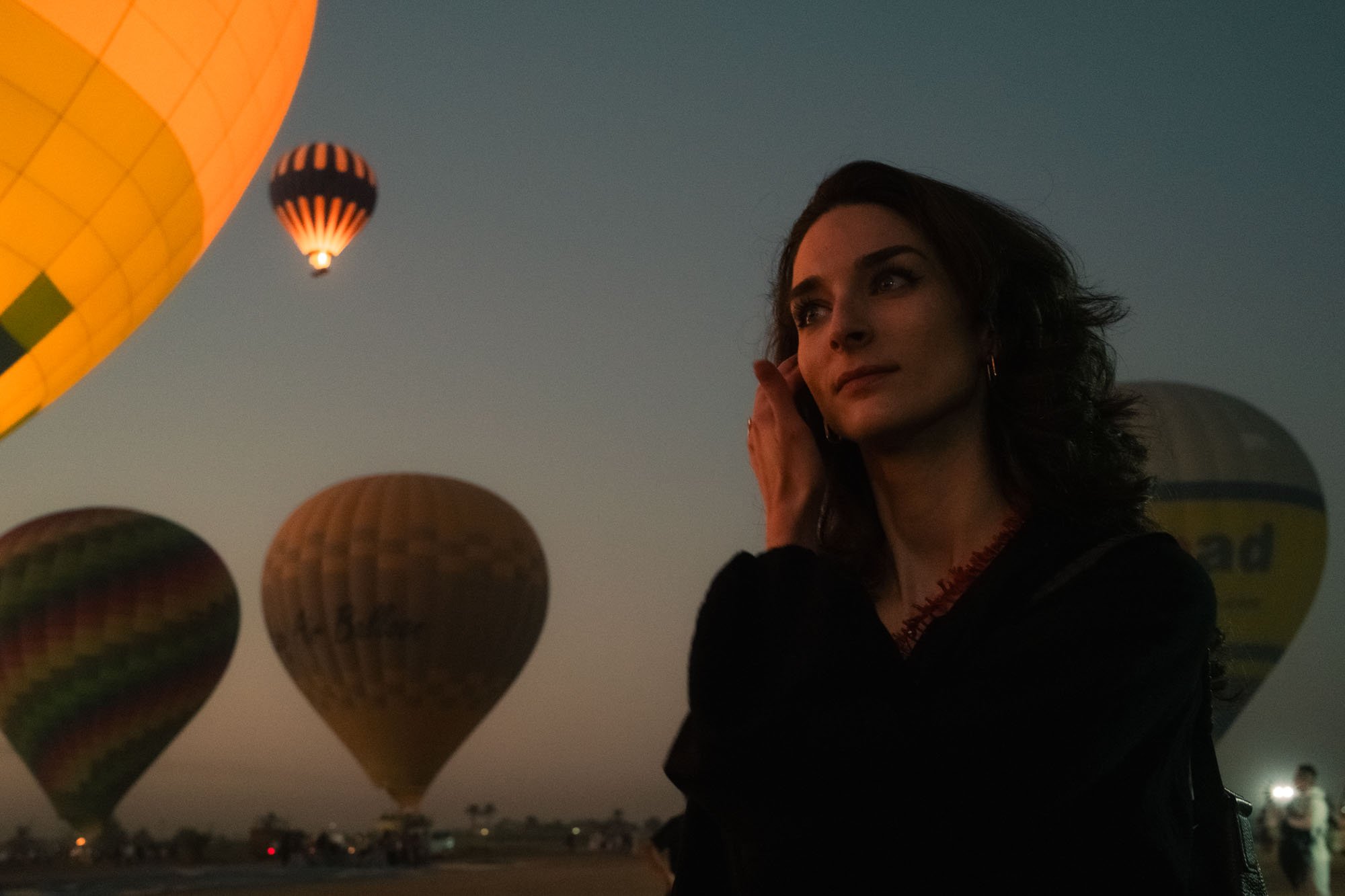 Une femme regarde vers le haut dans un ciel au crépuscule où flottent plusieurs montgolfières, l'une d'elles étant orange et deux autres avec des motifs colorés en egypte