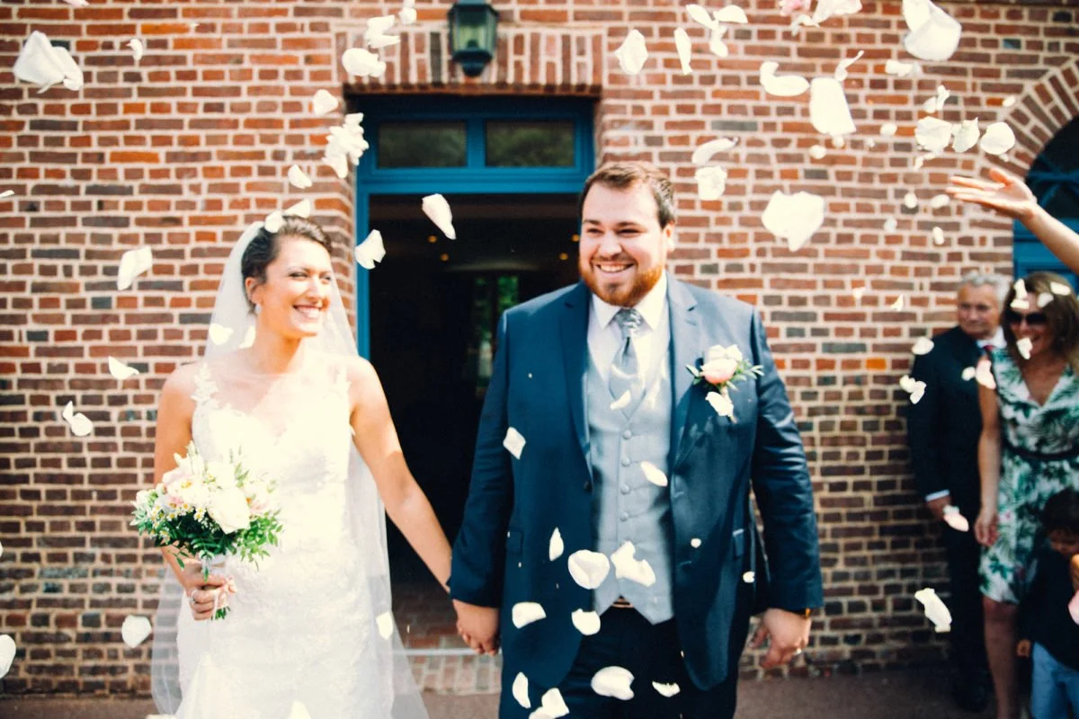 Un couple de mariés souriant tenant la main devant une maison en briques, entouré de fleurs et de confettis blancs, célébrant leur mariage.