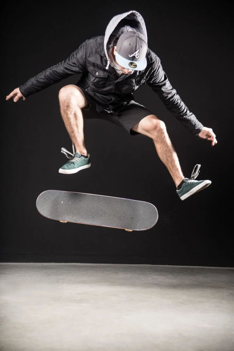 Joseph Garbaccio portant un casquette et un manteau noir effectuant un saut avec sa planche à skateboard dans un environnement intérieur avec fond noir.