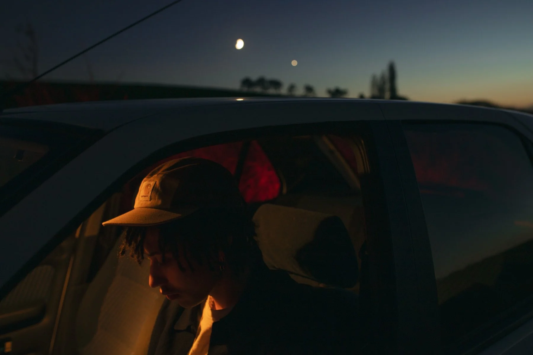 Une personne portant une casquette à l'intérieur d'une voiture, vue de l'extérieur au crépuscule, avec la lune et des étoiles visibles dans le ciel.