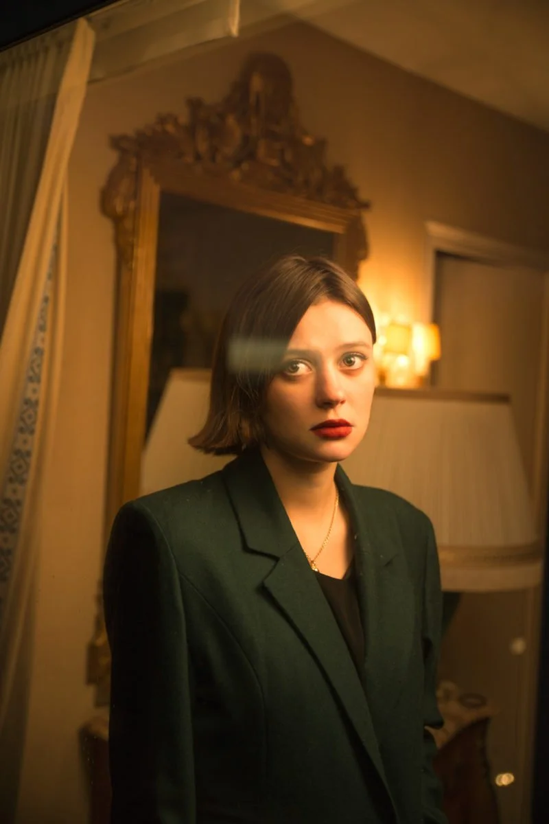 Une femme aux cheveux court et bruns, portant un blazer vert foncé, se tient dans une pièce élégante avec un miroir ancien en bois, un lampadaire et des rideaux dorés en arrière-plan.