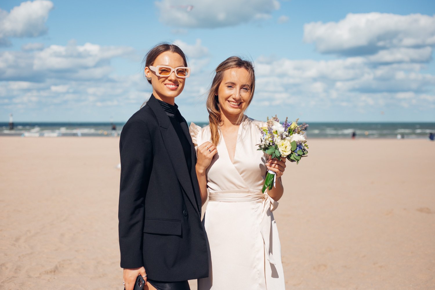 Deux femmes souriantes posant sur la plage, l'une tenant un bouquet de fleurs, un ciel partiellement nuageux en arrière-plan.