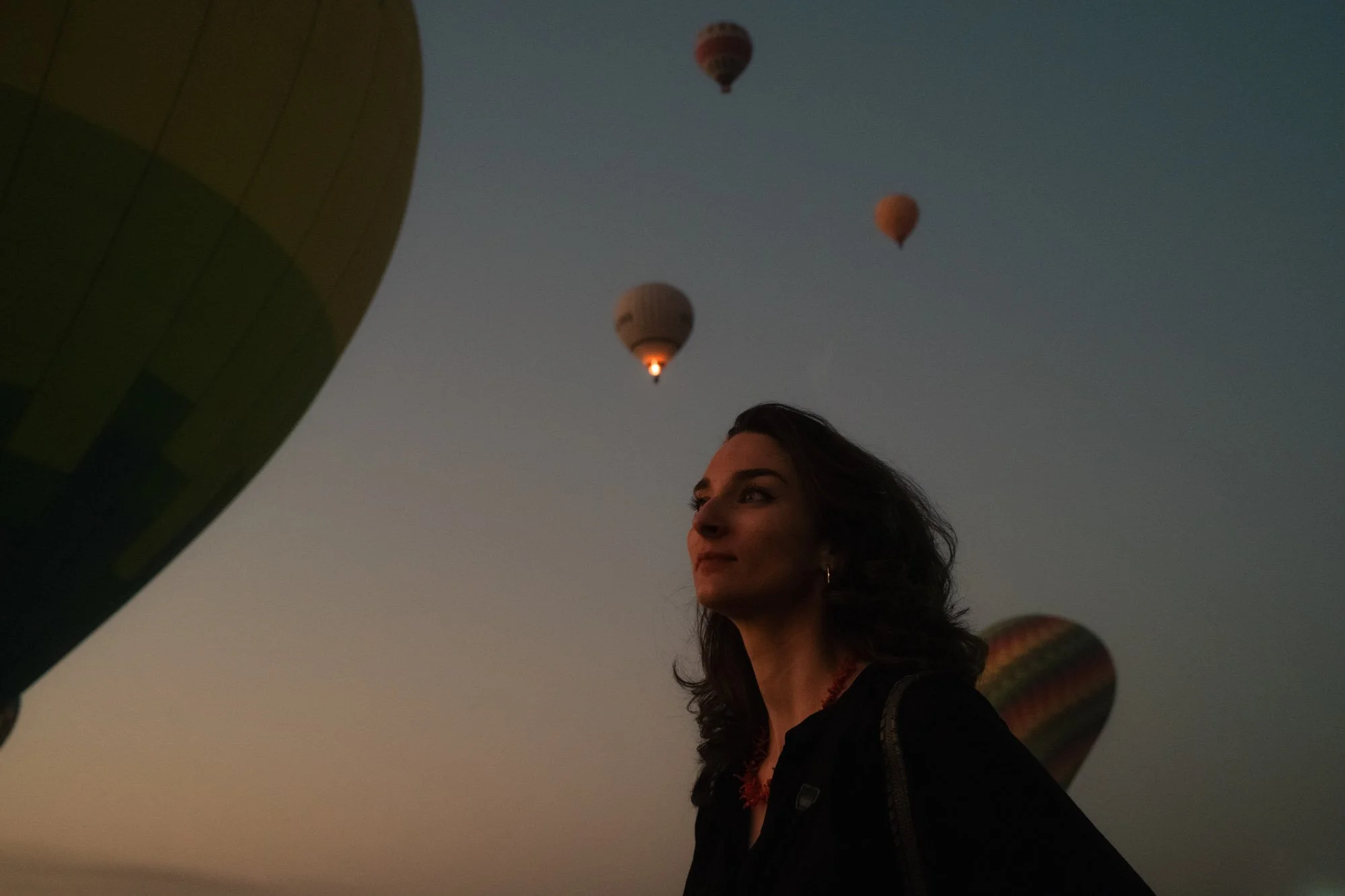 Une femme avec des cheveux longs et ondulés regarde vers le haut, dans un ciel crépusculaire, avec des montgolfières flottant à distance en egypte