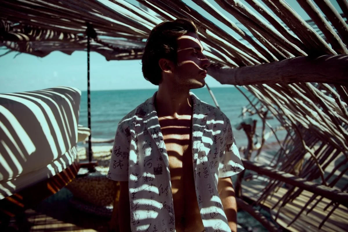 Un jeune homme debout sous un shelter en bambou au bord de la mer, avec des rayures de lumière et d'ombre sur lui, portant une chemise imprimée.