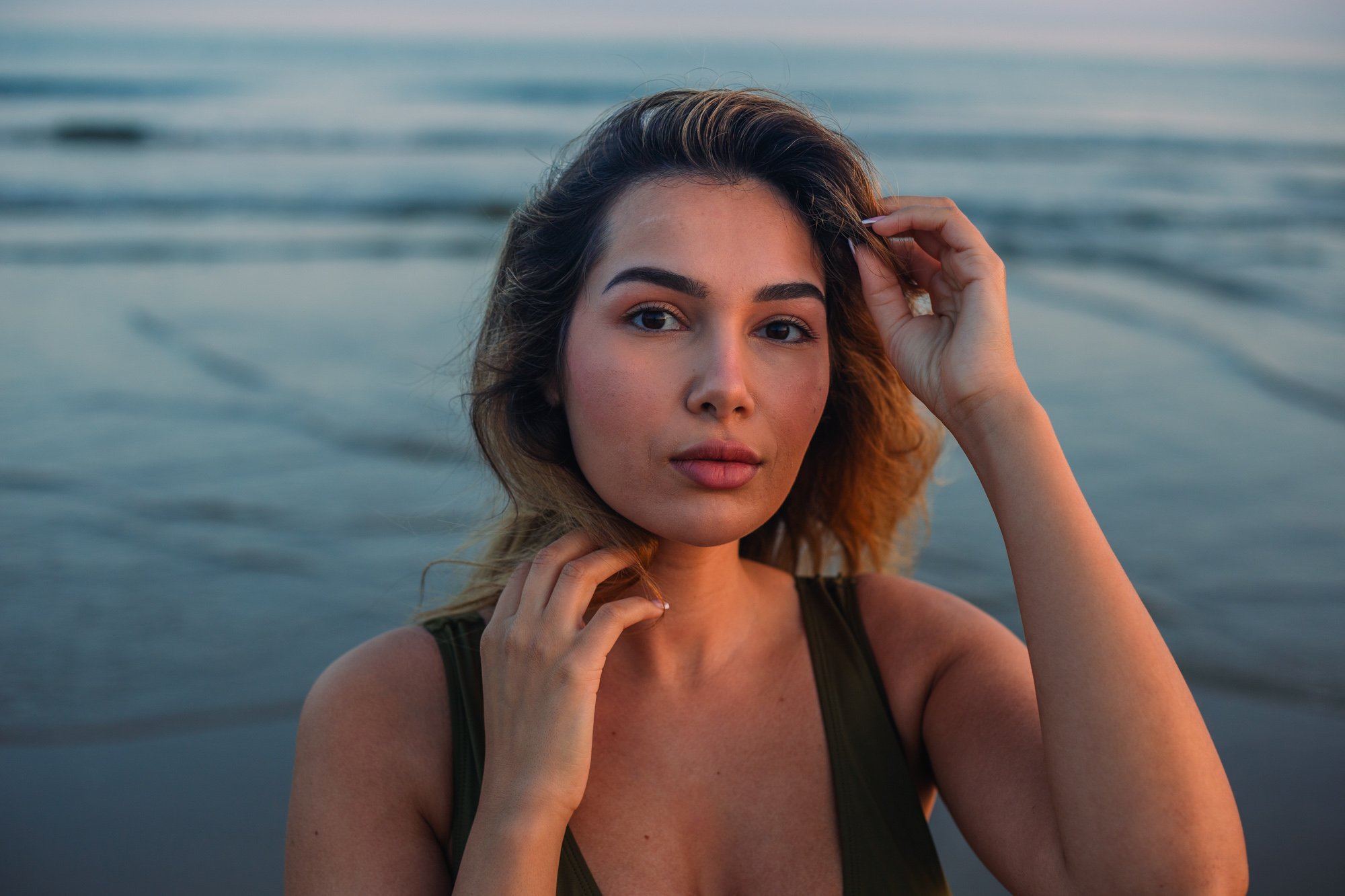 Jeune femme avec cheveux longs et yeux clairs, posant près de l'eau lors d'un coucher de soleil, portant un top noir, regardant la caméra.
