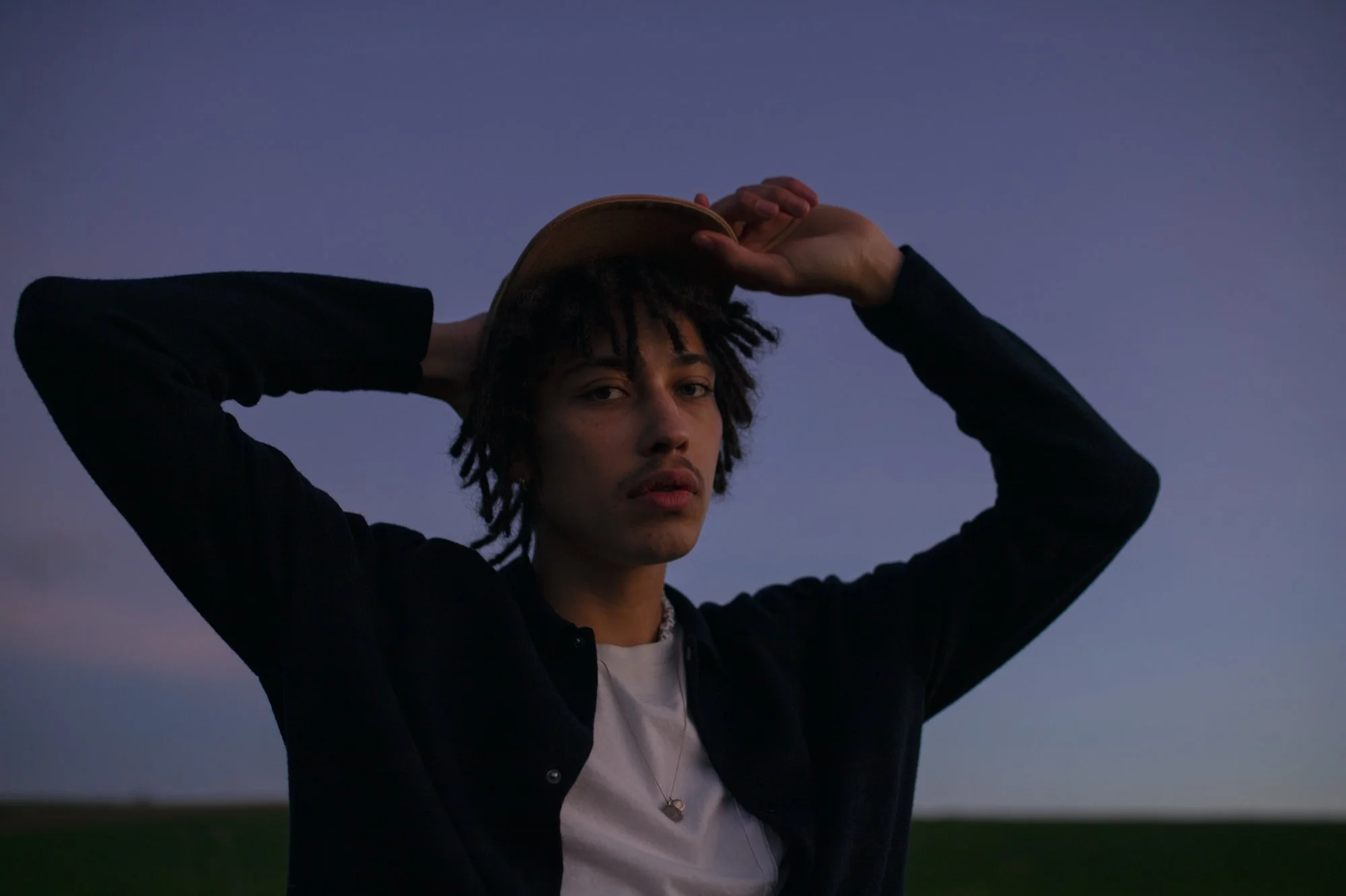 Jeune homme avec dreadlocks, portant une veste noire et un t-shirt blanc, tenant un chapeau au-dessus de la tête, sous un ciel en fin de journée ou crépuscule.