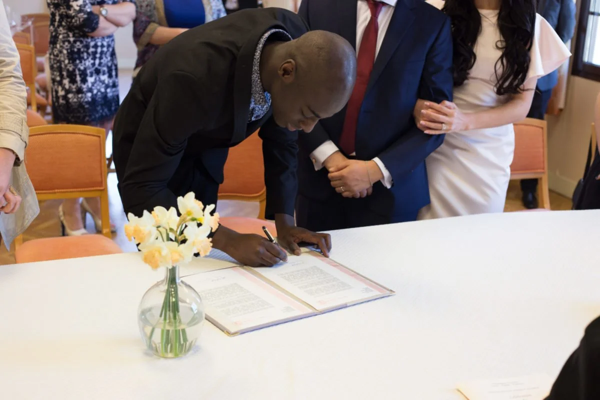 Une personne signe un document lors d'une cérémonie officielle, entourée de plusieurs personnes en costume et dans un cadre formel, avec une vase de fleurs blanches en premier plan.