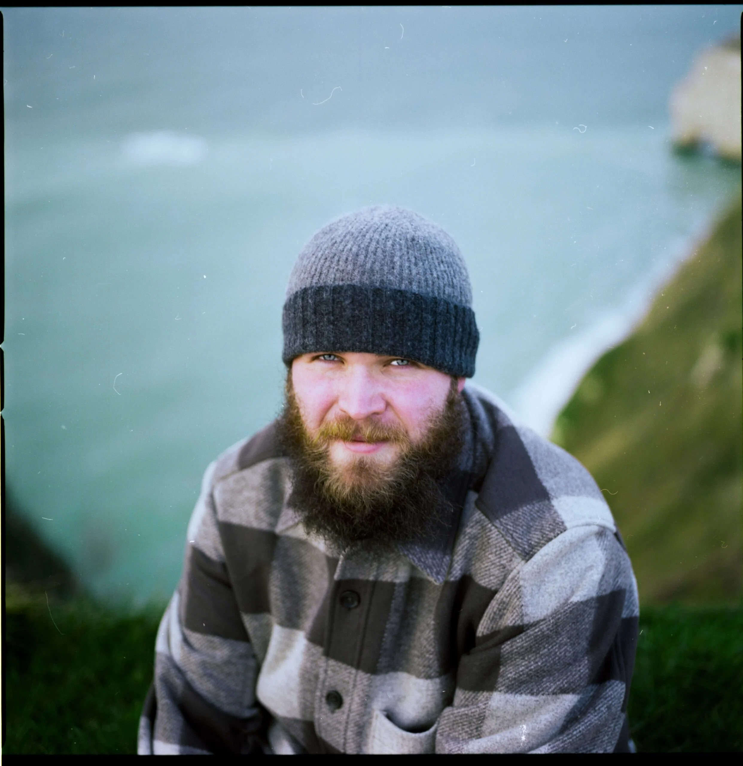 Jeune homme avec barbe, portant un bonnet en laine gris, vêtu d'un pull en laine gris à rayures et d'une chemise à carreaux, à l'extérieur, avec un fond flou de verdure.