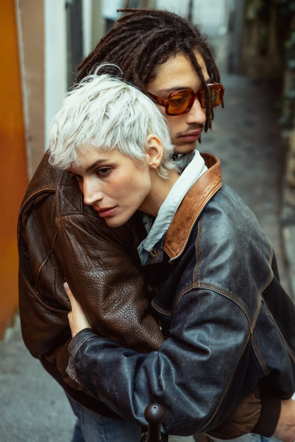 Deux personnes s'embrassent dans une rue, l'une avec des cheveux blonds courts, l'autre avec des dreadlocks et des lunettes de soleil.