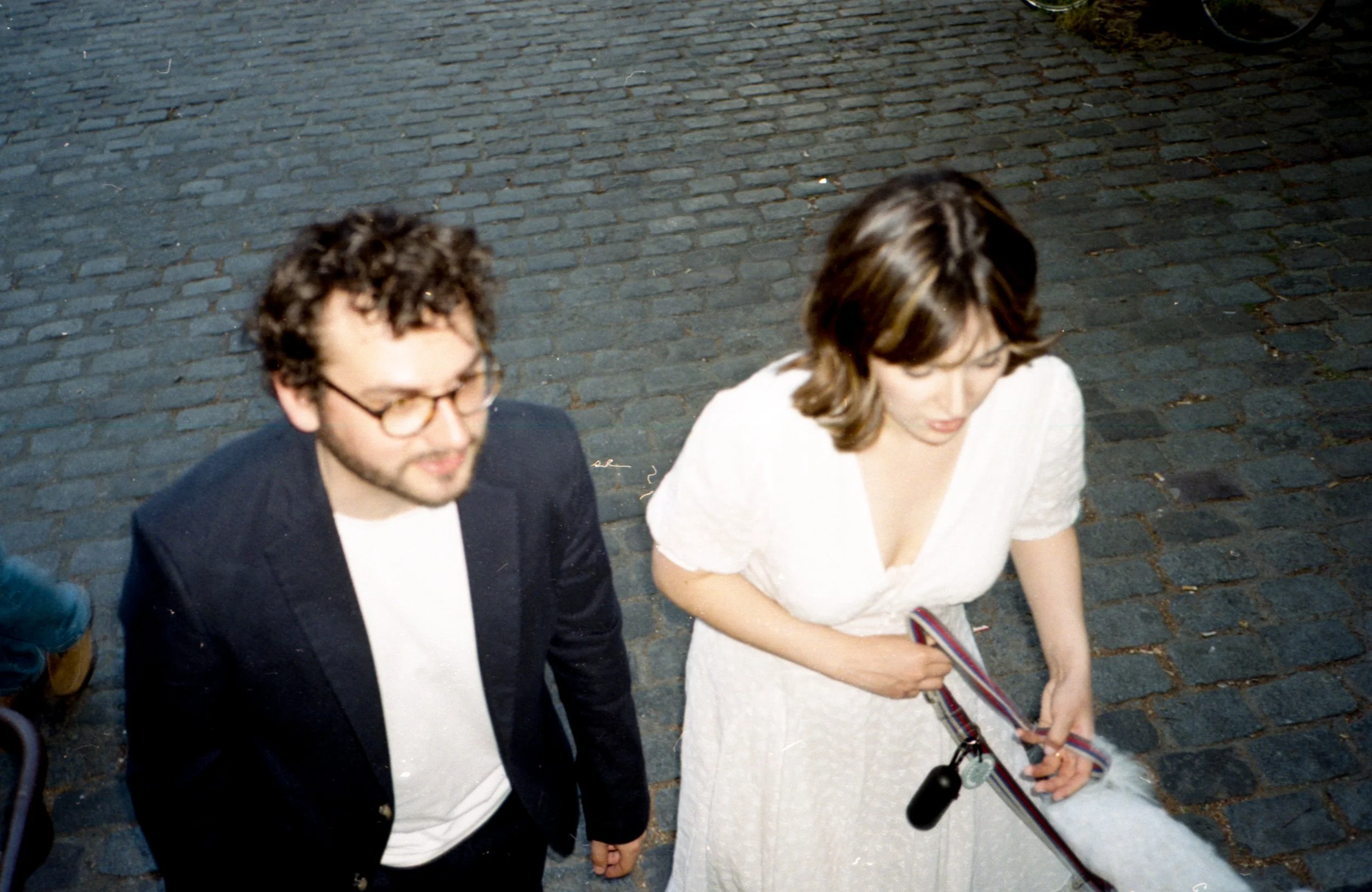 Deux mariés assises côte à côte sur un trottoir pavé, une femme en robe blanche avec une chaîne de clés à la main, un homme en veston noir avec des lunettes.