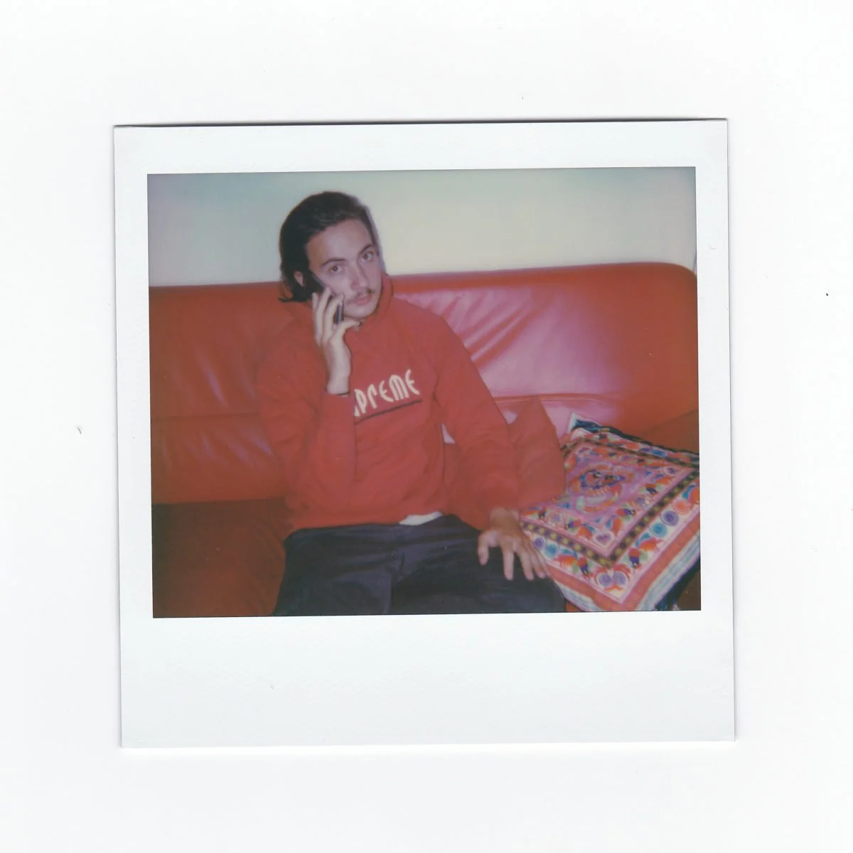 Lord Esperanza assis sur un canapé rouge parlant au téléphone, portant un sweat rouge et un pantalon noir, avec un coussin coloré à côté.