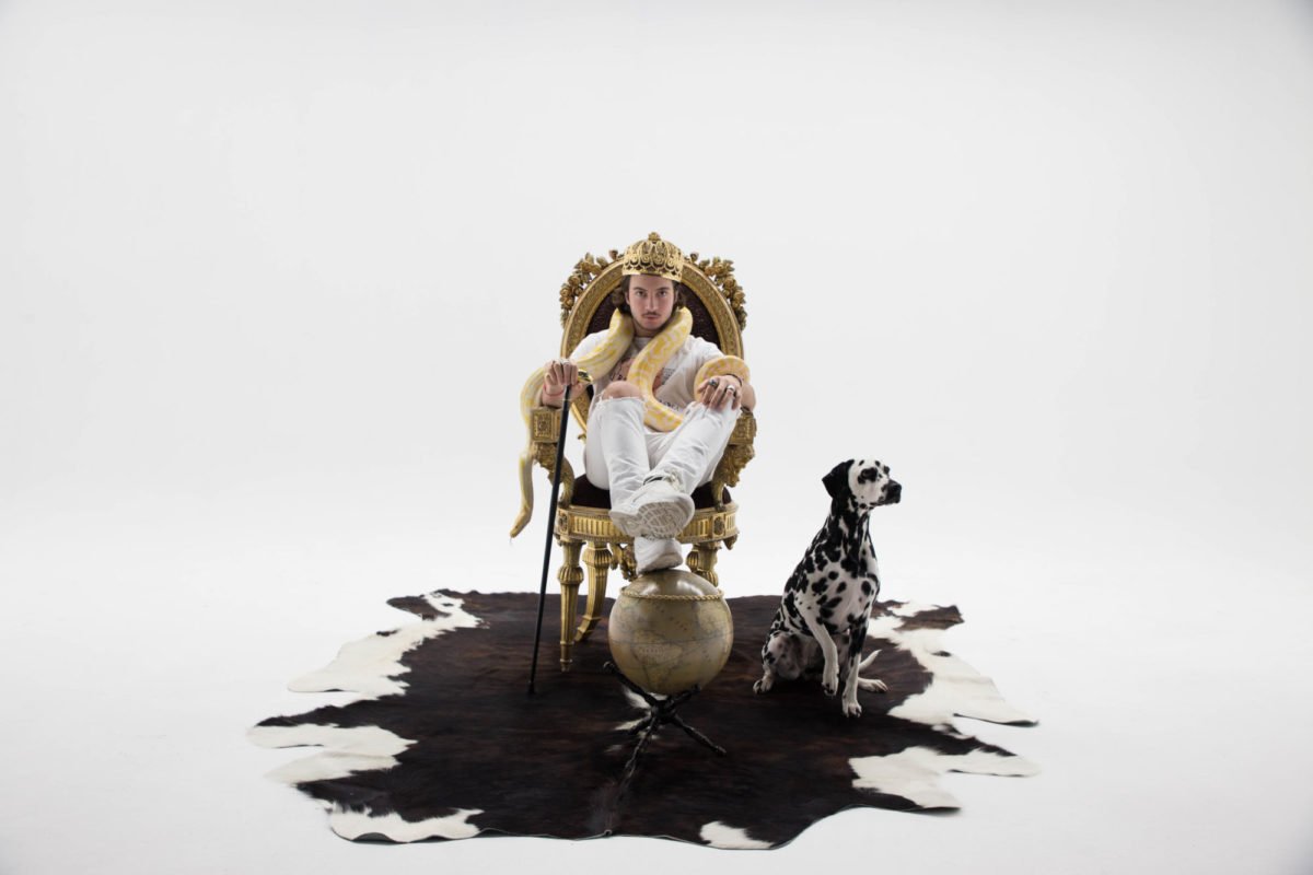 Un homme assis sur un trône doré avec une couronne, une serpente jaune autour du cou, une canne à main, et une vieille planète en dessous de la chaise. À côté de lui, un Dalmatien assis sur une peau de vache blanche et noire. L'ensemble est disposé s