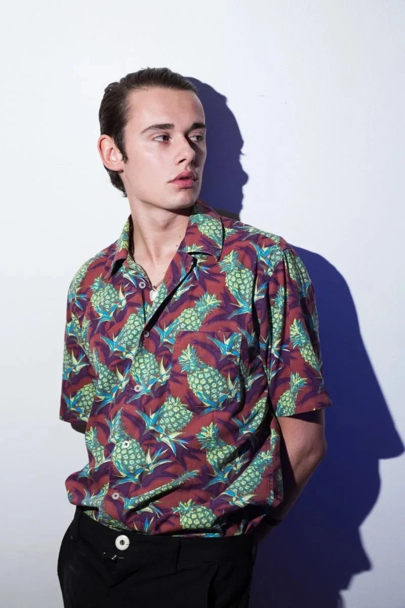 Un jeune homme portant une chemise imprimée d'ananas, debout contre un mur blanc, avec un ombrage marqué derrière lui.