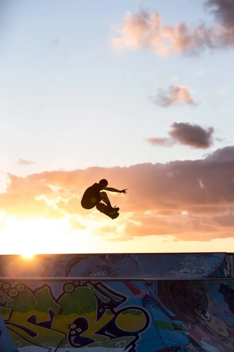 Un skatepark au coucher du soleil avec un skateur en l'air, silhouette noire, et des graffitis colorés sur la rampe.