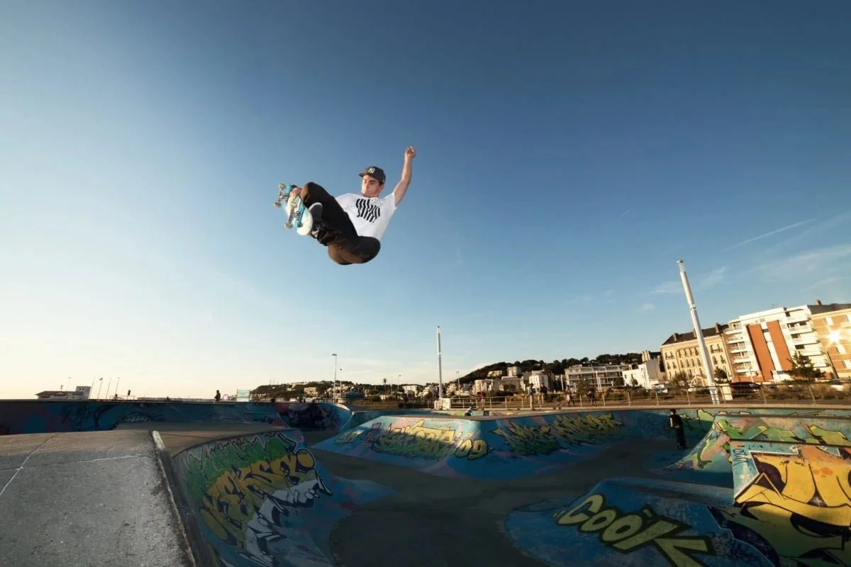 Joseph Garbaccio en plein saut dans un skatepark avec un ciel clair et des bâtiments en arrière-plan.