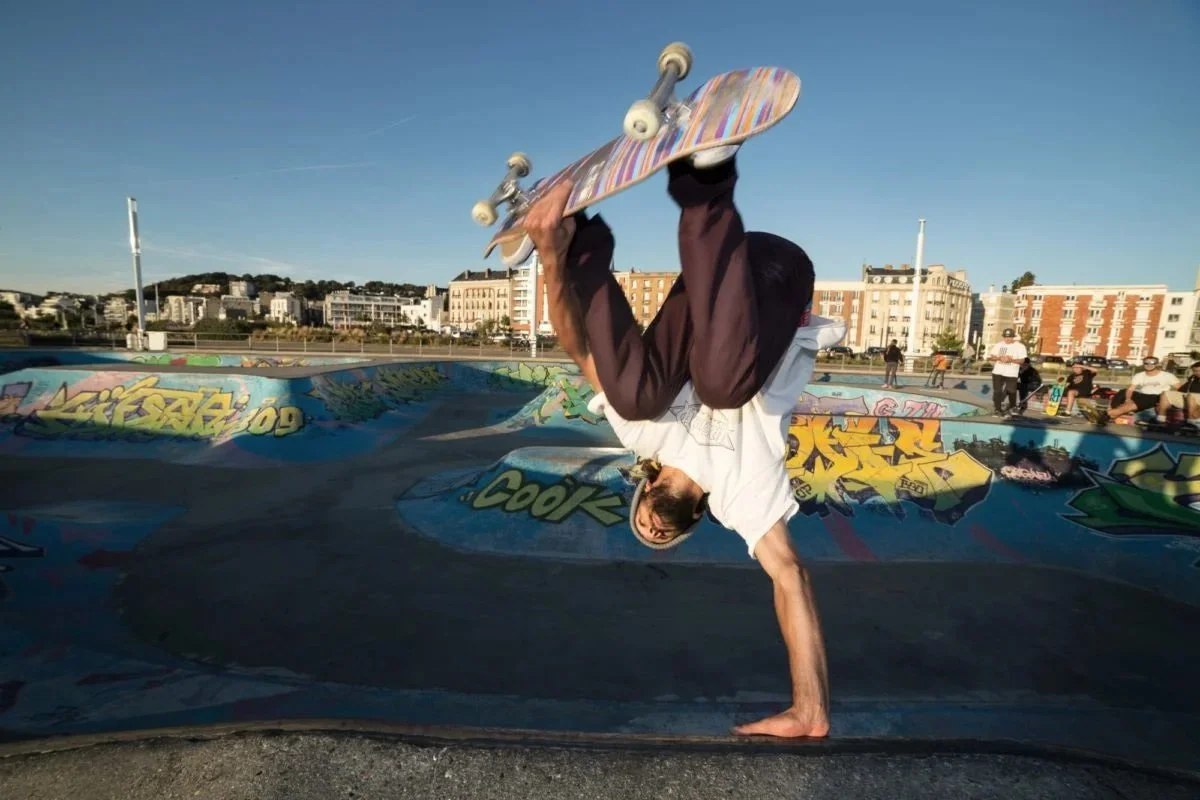 Un skateur effectuant une figure sur un bowl de skatepark avec graffiti, en plein air, avec des bâtiments en arrière-plan et un ciel clair.