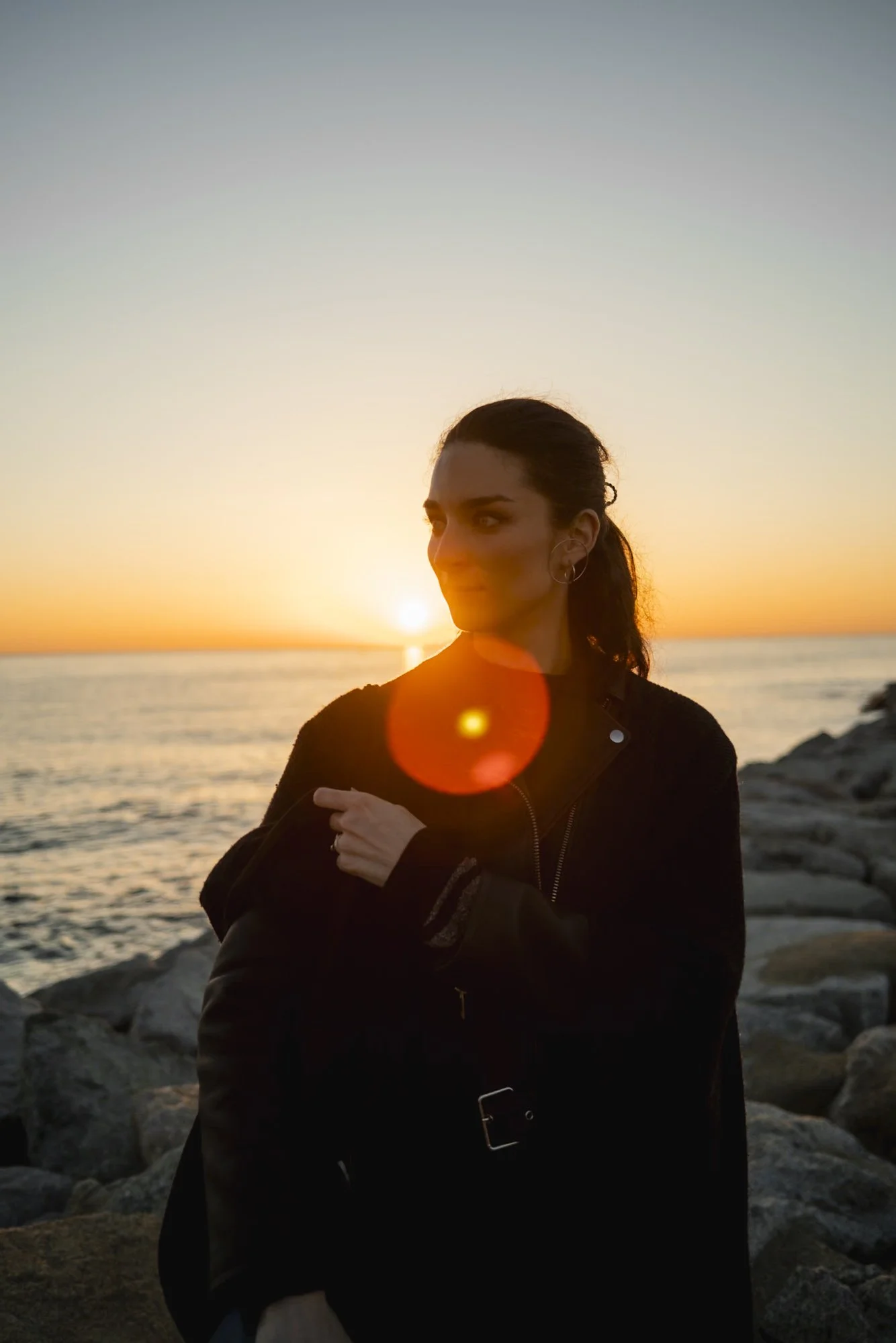 Une femme debout sur les rochers près de la mer au coucher du soleil, regardant à gauche, portant une veste noire, avec la lumière du soleil créant une lentille sur l'image.