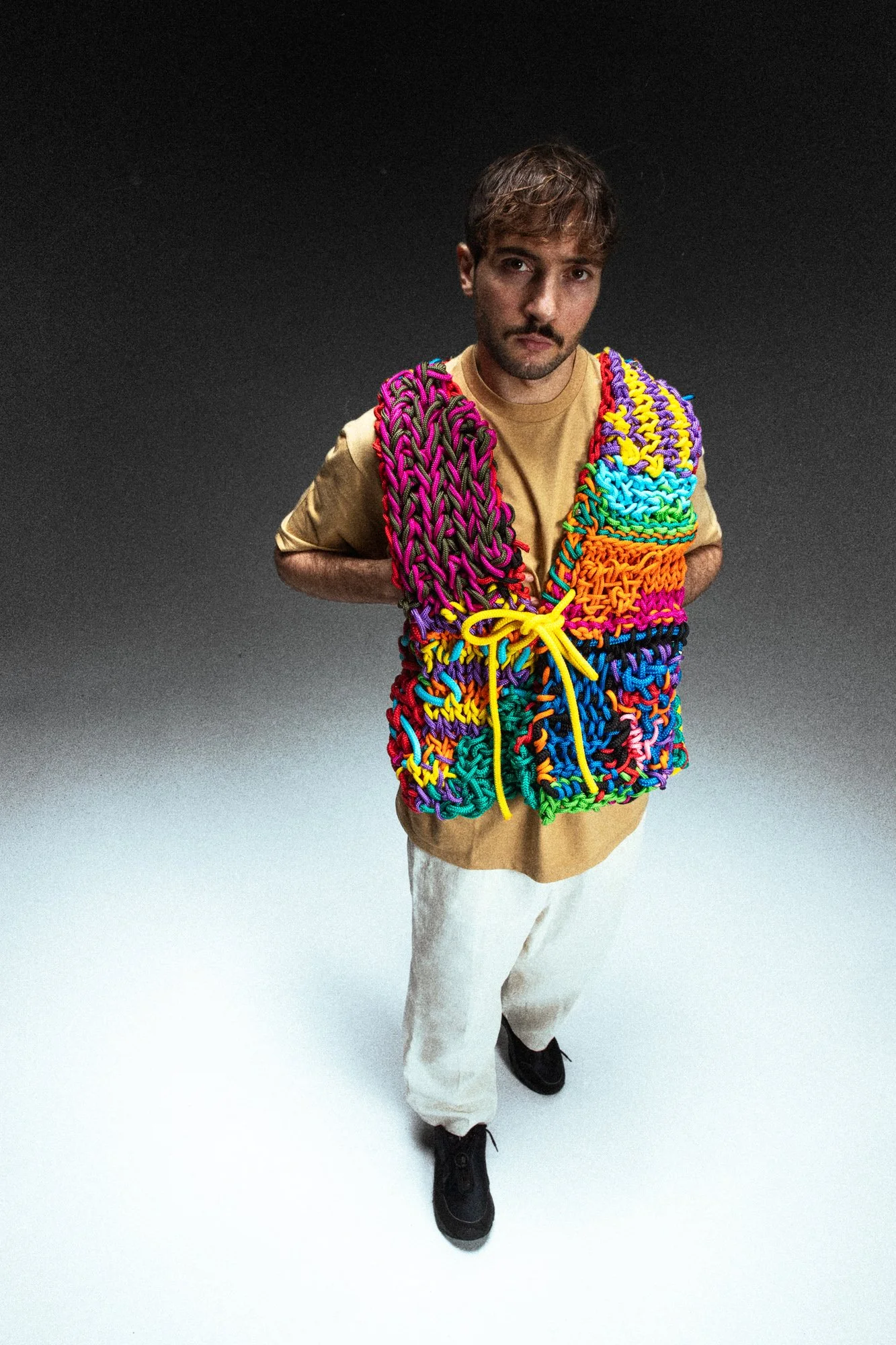 Un jeune homme porte un sac en filet fait de cordages colorés, avec des couleurs vives, sur un fond sombre.