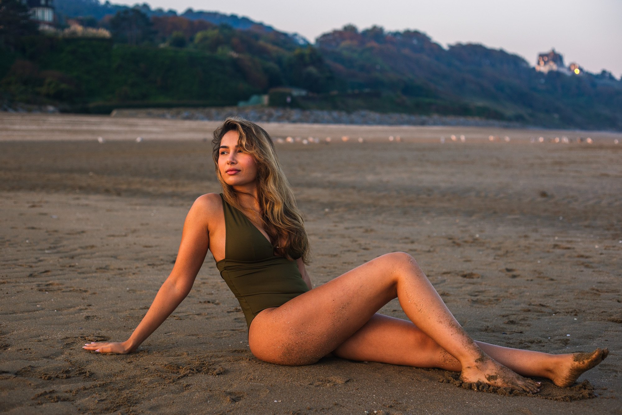 Jeune femme en maillot confortable orange foncé, assise sur la plage de sable, regardant vers la gauche avec un paysage arrière comprenant des collines et un château en haut, le soir ou au coucher du soleil.