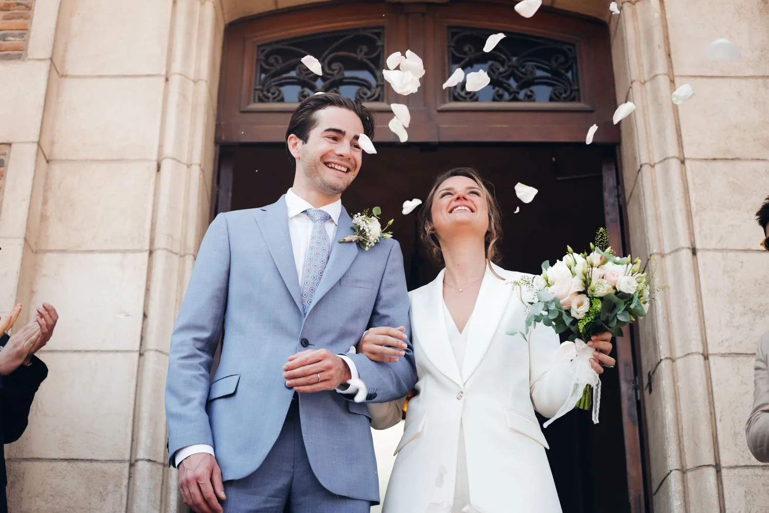 Un couple de mariés souriants sortant de l'église, la femme tenant un bouquet de fleurs, des pétales de rose tombant autour d'eux, célébrant leur mariage.