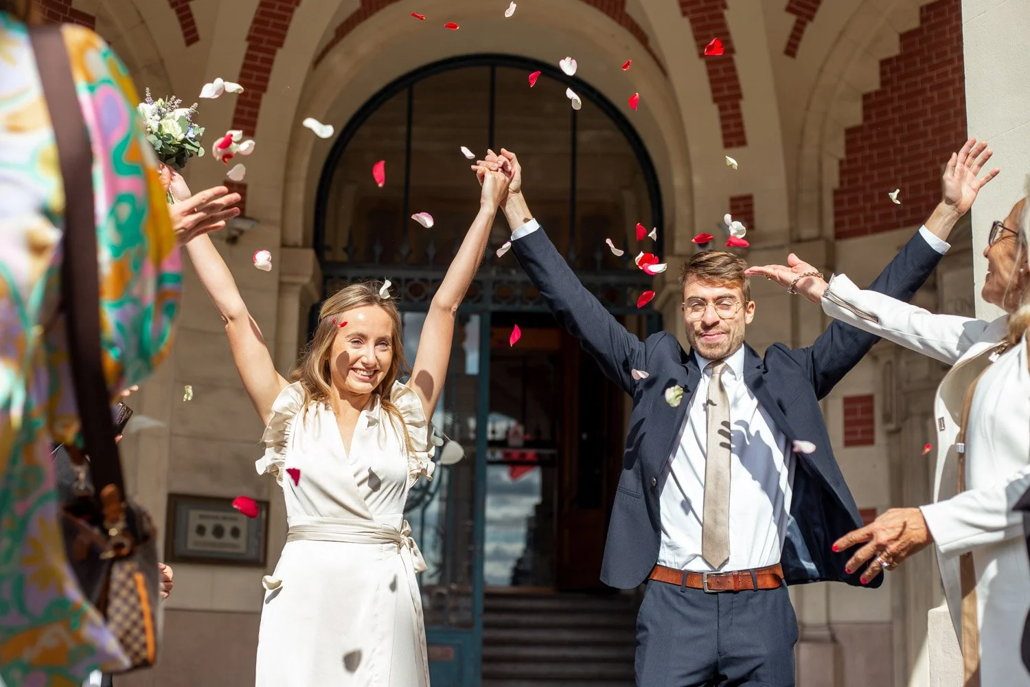 Un couple de mariés célébrant leur mariage devant une entrée en pierre, entourés de convives qui lancent des confettis colorés.