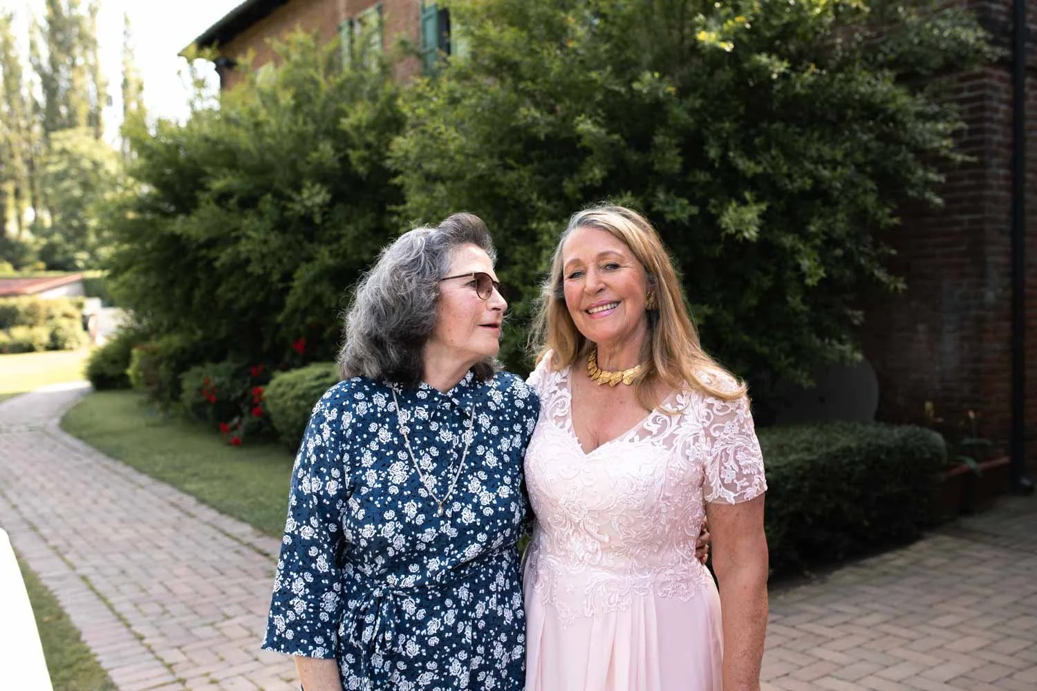 Deux femmes souriantes posent ensemble à l'extérieur, dans un jardin verdoyant avec une allée en pavés, une derrière une grande haie et une autre près d'une bâtisse en briques, en tenue élégante.