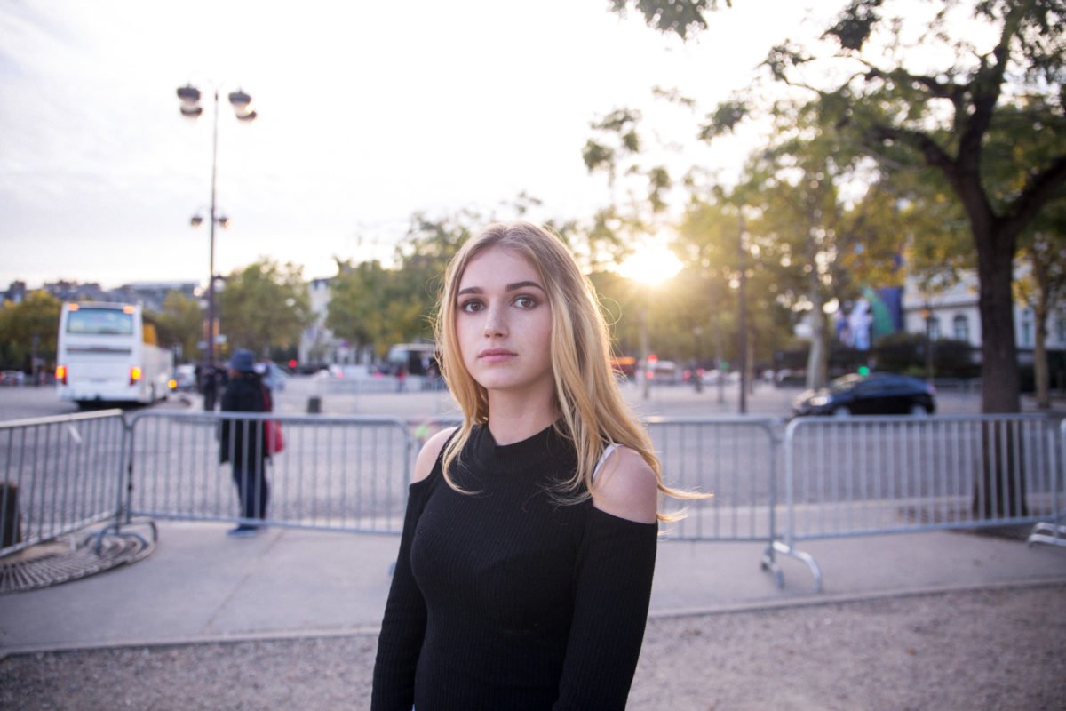 Jeune femme avec des cheveux blonds au coucher du soleil dans une rue, portant une robe noire à épaules dénudées, devant une barrière.