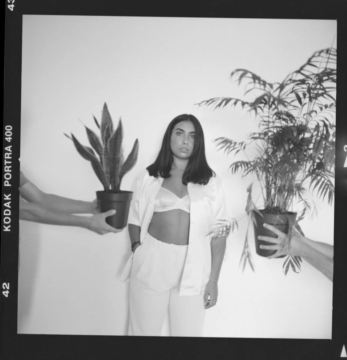 Une jeune femme en tenue décontractée pose devant un mur blanc, entourée de deux personnes dont on voit seulement les bras, chacune tenant un pot de plante.