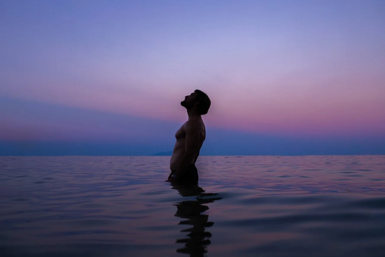 Un homme torse nu, debout dans l'eau, regarde vers le ciel au coucher du soleil ou à l'aube, avec des couleurs de pastel dans le ciel et une mer calme.
