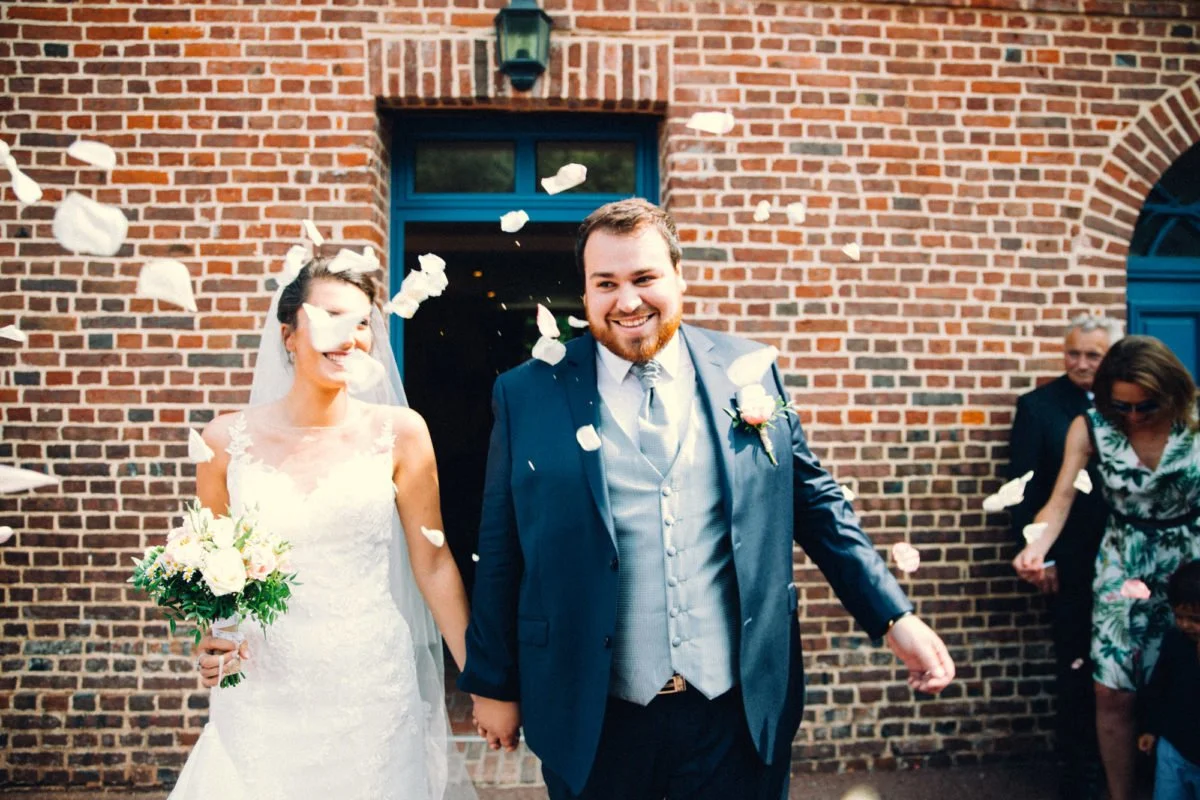 Un jeune couple de mariés souriants sortant d'une cérémonie religieuse, la mariée tenant un bouquet de fleurs, des pétales de roses sont jetés en l'air par les invités contre un mur en briques.