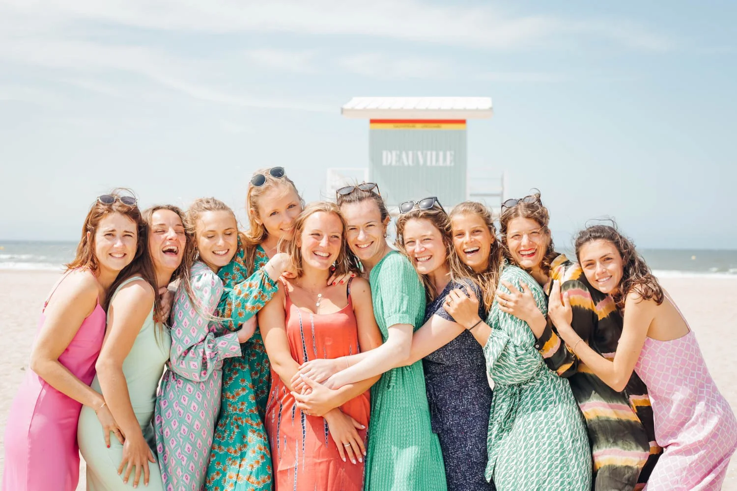Groupe de femmes souriantes en robes colorées sur la plage avec un ciel bleu et la mer à l'arrière-plan. Se trouve un panneau avec l'inscription "Deauville".