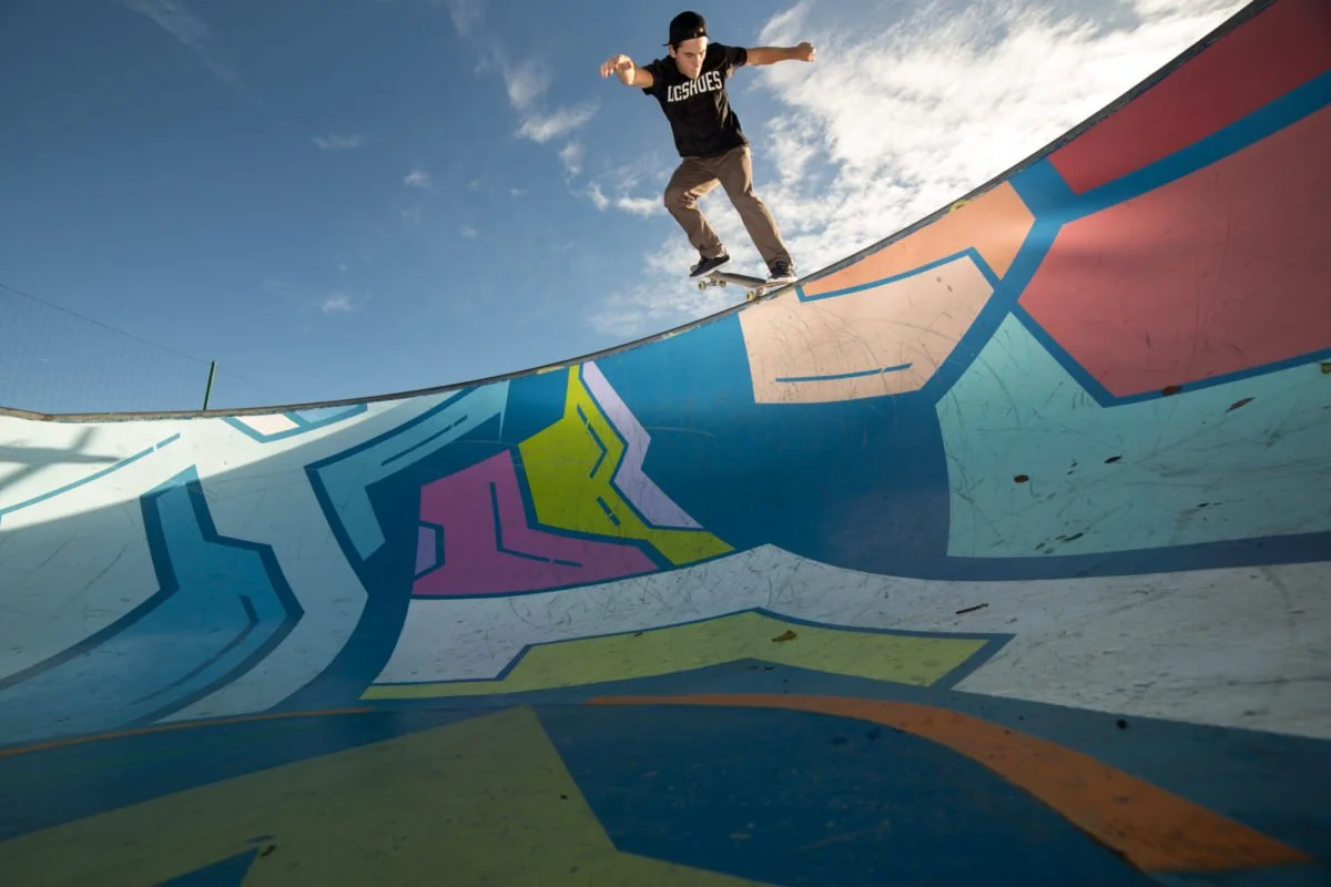 Joseph Garbaccio en skateboard en train de faire une figure dans un skatepark en béton peint avec des formes et des couleurs vives, sous un ciel clair avec quelques nuages.