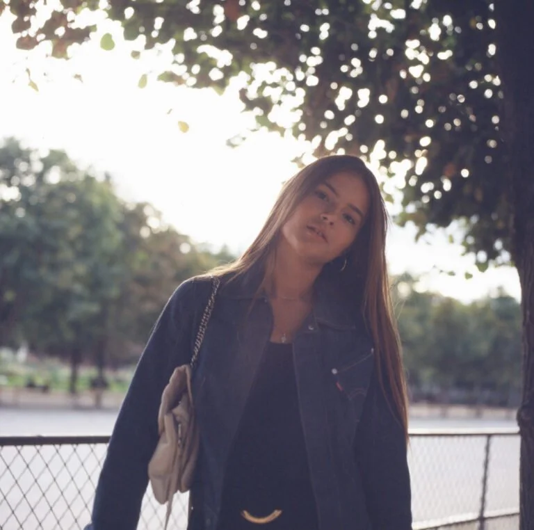 Jeune femme avec long cheveux, portant une veste en denim et un sac beige, debout à côté d'un arbre dans un parc, avec un éclairage naturel en arrière-plan.