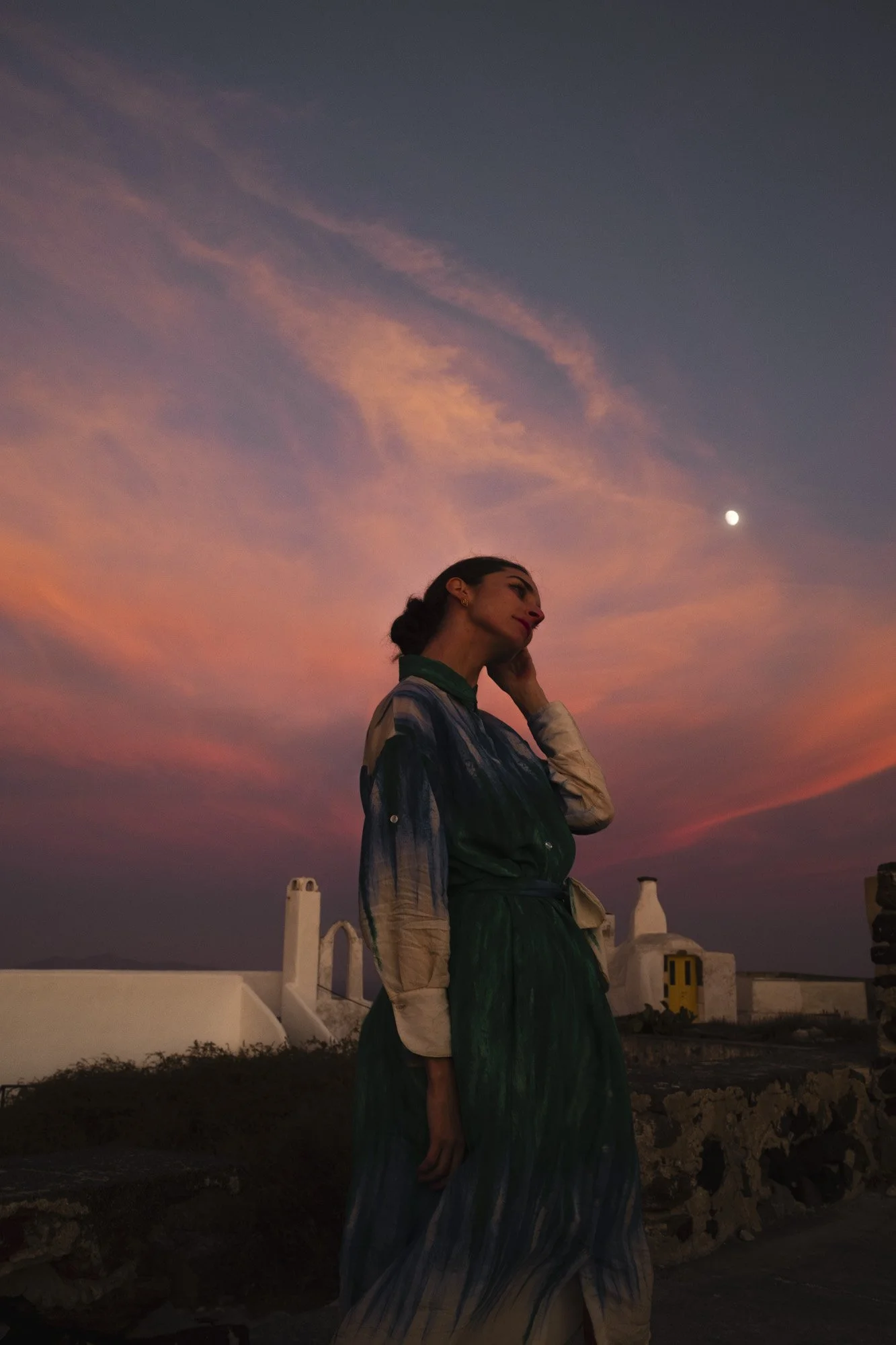 Une femme debout à côté d'une clôture en pierre, regardant vers la droite, avec un ciel coloré au coucher du soleil et la lune visible en arrière-plan à santorin