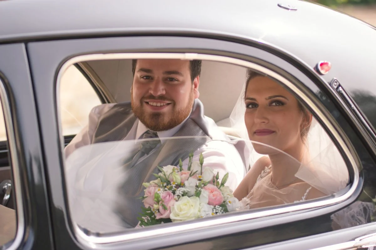 Un couple de mariage dans une voiture noire, la mariée tient un bouquet de fleurs. L'homme porte un costume gris, la femme un voile et une robe blanche.
