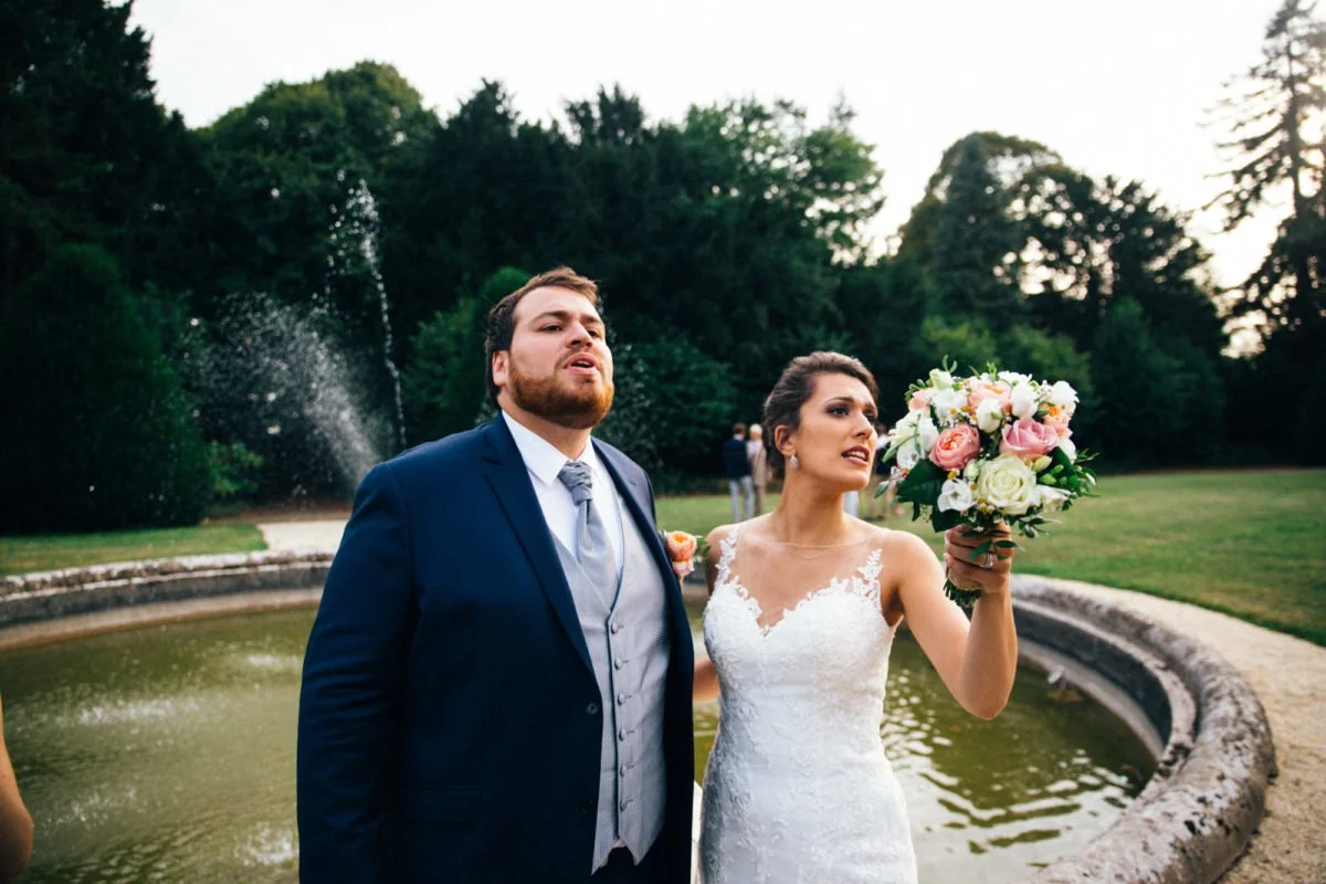 Un couple lors d'un mariage en extérieur, la femme avec une robe blanche et un bouquet, l'homme en costume bleu, près d'une fontaine dans un parc.