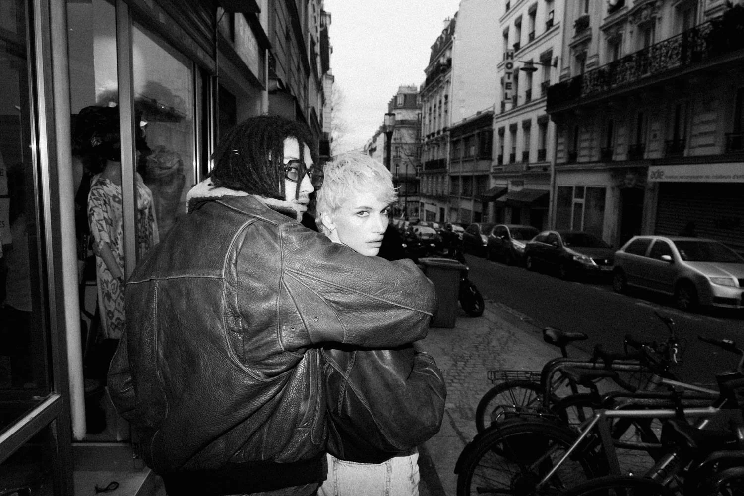 Deux personnes regardent vers la caméra dans une rue urbaine, avec des voitures garées et des bâtiments en arrière-plan, au coucher du soleil.