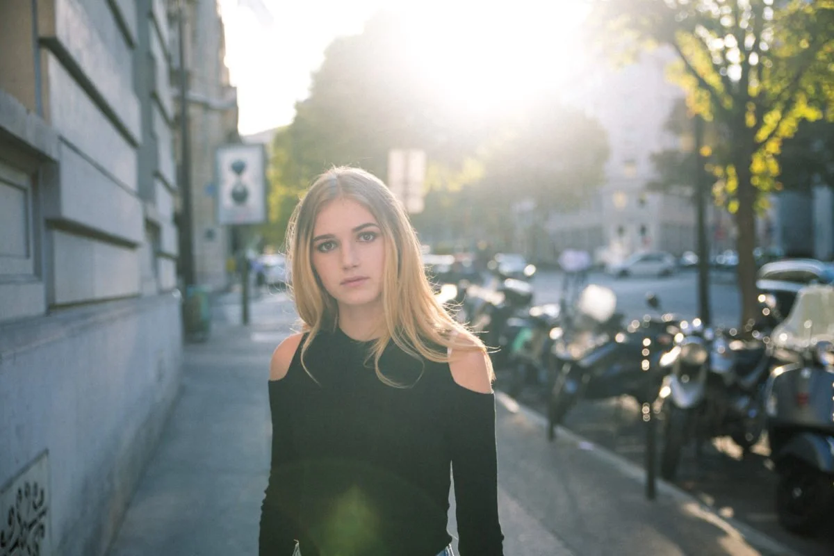 Jeune femme au cheveux longs, portant un pull noir avec des épaules découpées, qui se tient sur un trottoir en ville, avec des motos et des voitures en arrière-plan, sous une forte lumière solaire