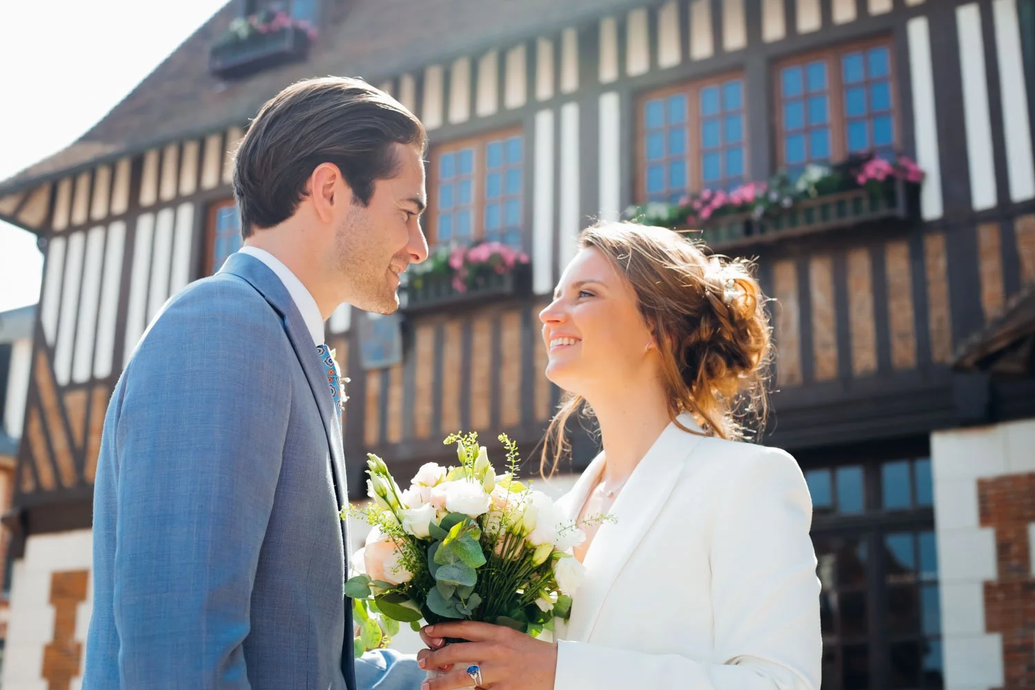 Un couple qui se regarde tendrement, la femme tient un bouquet de fleurs, devant une maison à colombages.